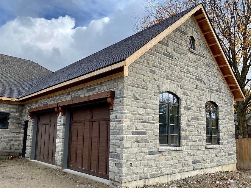 New detached garage clad in light stone with dark arched windows and wood doors
