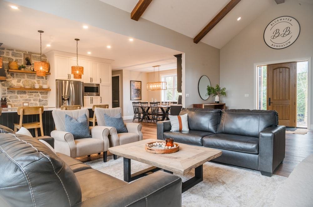 Open-plan great room with white kitchen, dark island, stone fireplace, and timber beams
